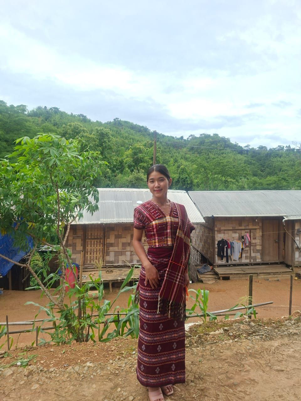Young woman in traditional Chin dress, refugee camp behind her